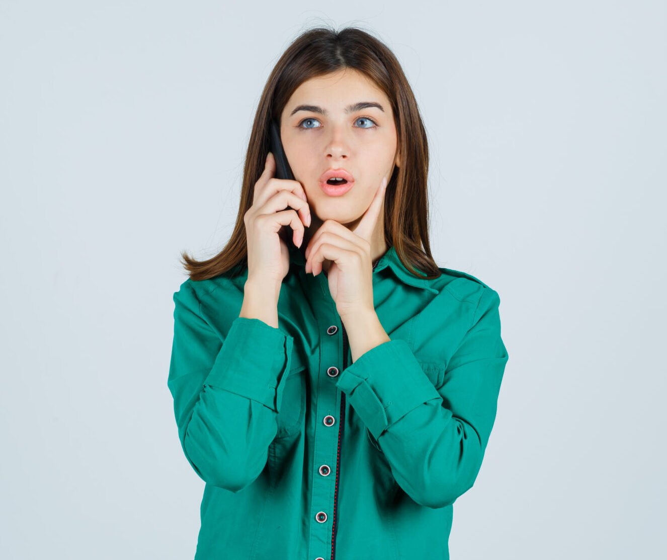 young girl talking to phone, putting index finger under chin in green blouse, black pants and looking surprised. front view.